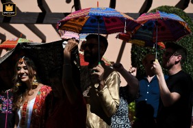 Une belle mariée, vêtue d'une tenue traditionnelle, entre gracieusement dans la cérémonie Haldi en plein air à l'Elba Estepona Gran Hotel & Thalasso Spa, entourée de femmes portant des parasols sous le chaud soleil.